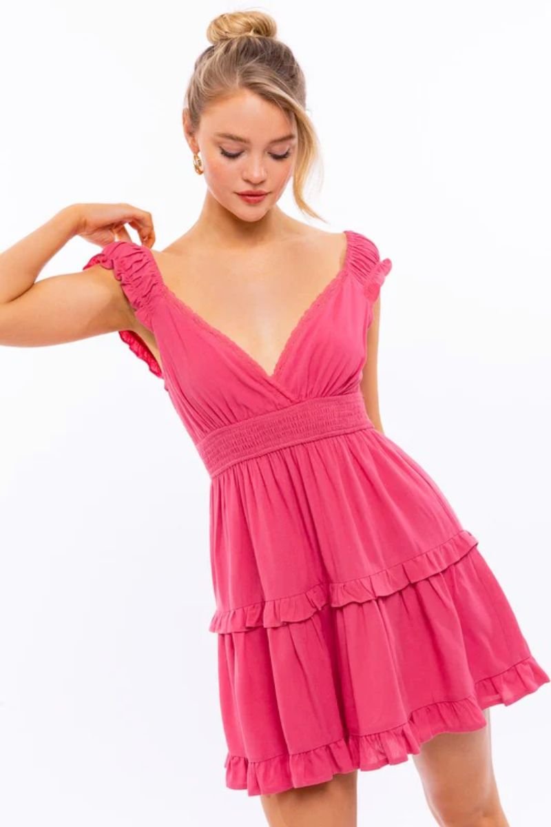 Cap Sleeve Ruffle Mini Dress. The model is wearing a pink frilly dress with a deep v neckline and her blonde hair is in a bun.