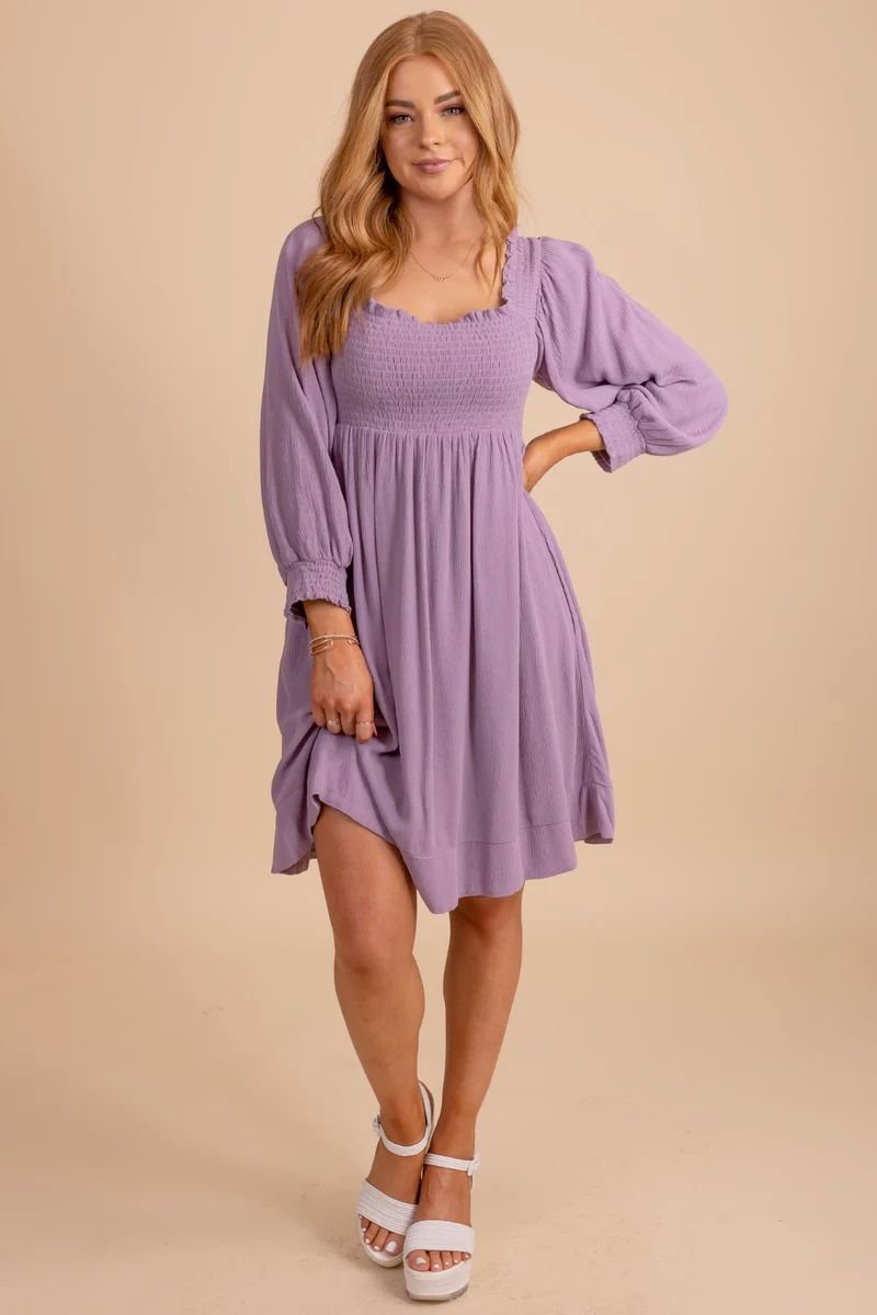 Gentle Gleam Long Sleeve Mini Dress. The model is wearing a purple dress with a ruched bodice and cuffed sleeves. It ends at the knees and she is wearing white heels.