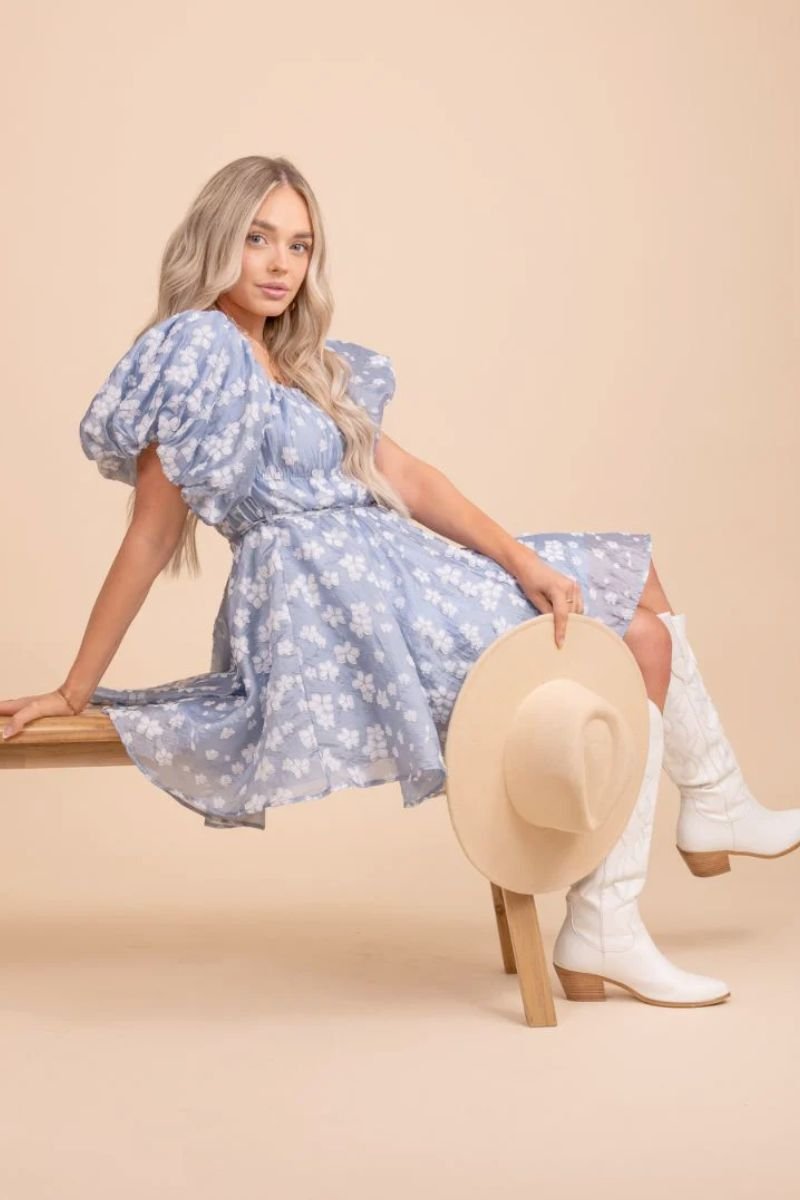Everything She Wants Mini Dress. The model is wearing a blue mini dress with white flowers on it and lots of extra fabric that makes a full, puffy skirt. The sleeves are puffy and short and she is holding a tan wide brimmed hat and white cowboy boots. She is sitting on a bench.