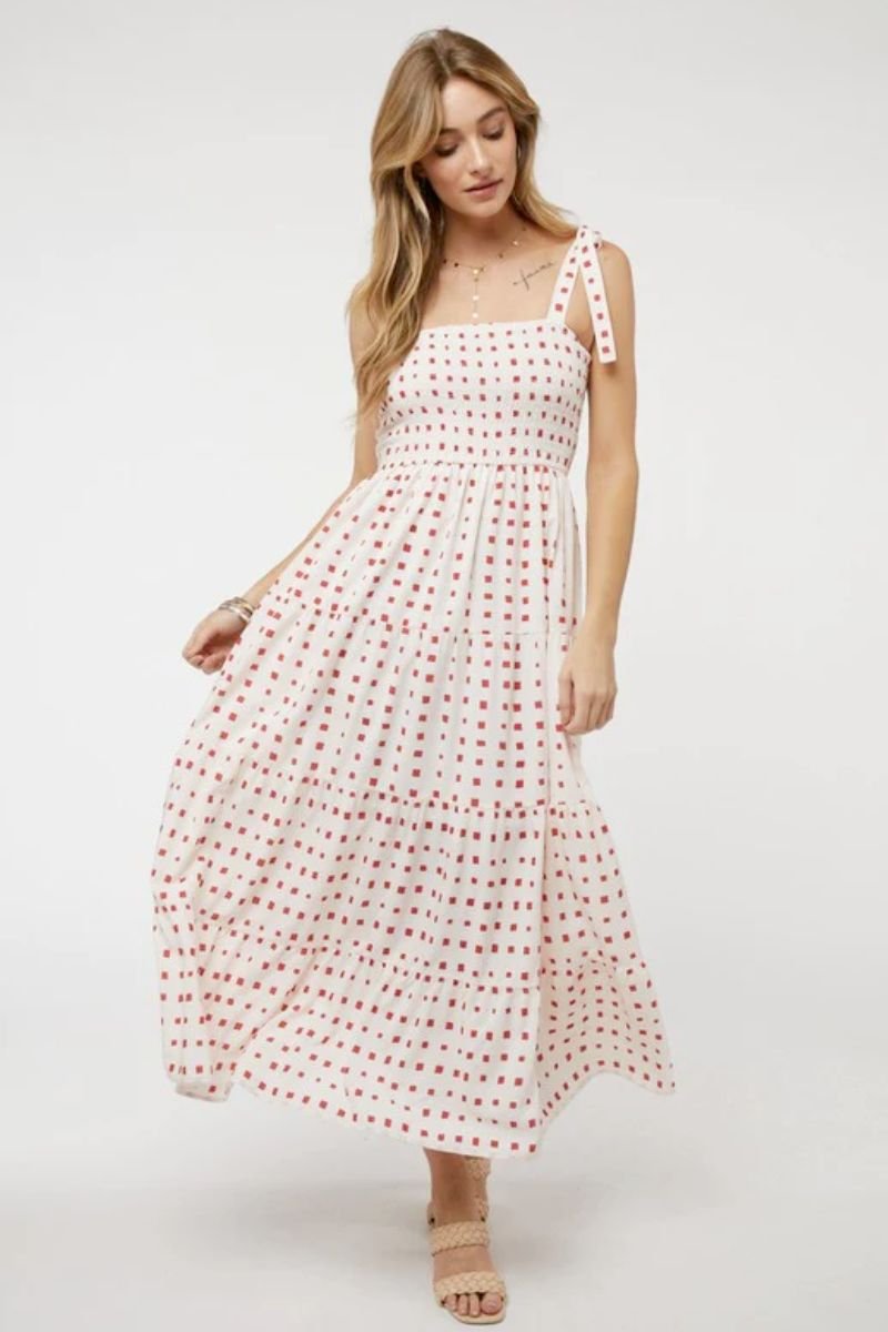 Printed Smocked Ruffle Maxi Dress. The model is wearing a white maxi dress with a red square printed pattern. There are shoulder ties for straps and the model is wearing tan heels.