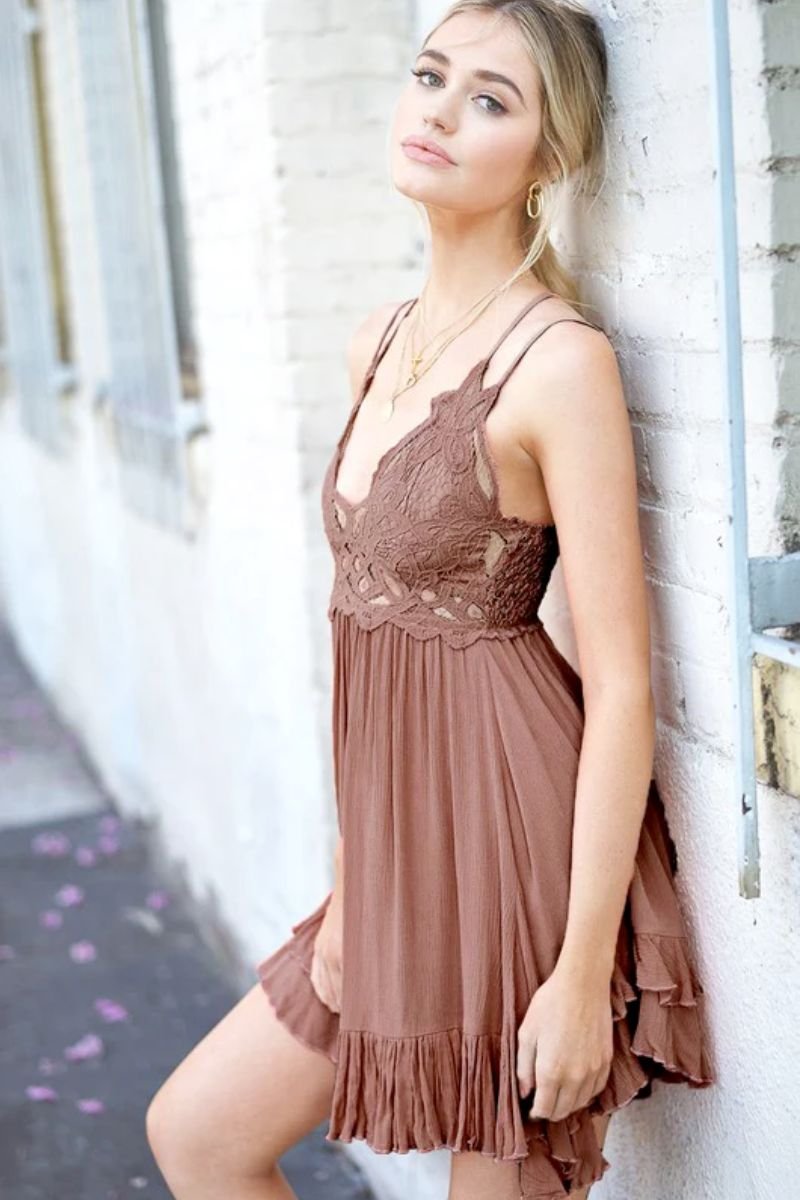 Monterey Dress. The model is wearing a brown mini dress with a flowing ruffled skirt and a lace bodice. The model is wearing gold jewelry and is outside a brick building.