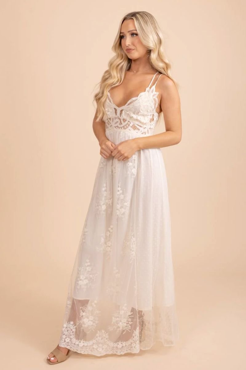 Dreamy Daze Lace Maxi Dress. The model is wearing an off white lace dress with a flowing silhouette and crochet top. The straps are also crossed in the back.