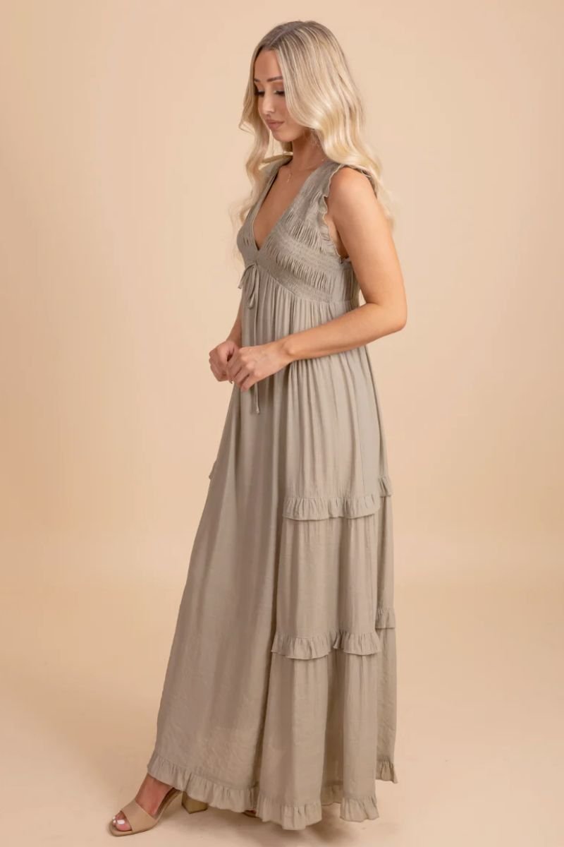 Now Until Forever V-Neck Maxi Dress. The model is wearing a light green floor length dress with a ruched bodice with a bow. There are no sleeves but two thick straps that are ruched. There are ruffles down the length of the dress and at the bottom and the model is also wearing tan heels.