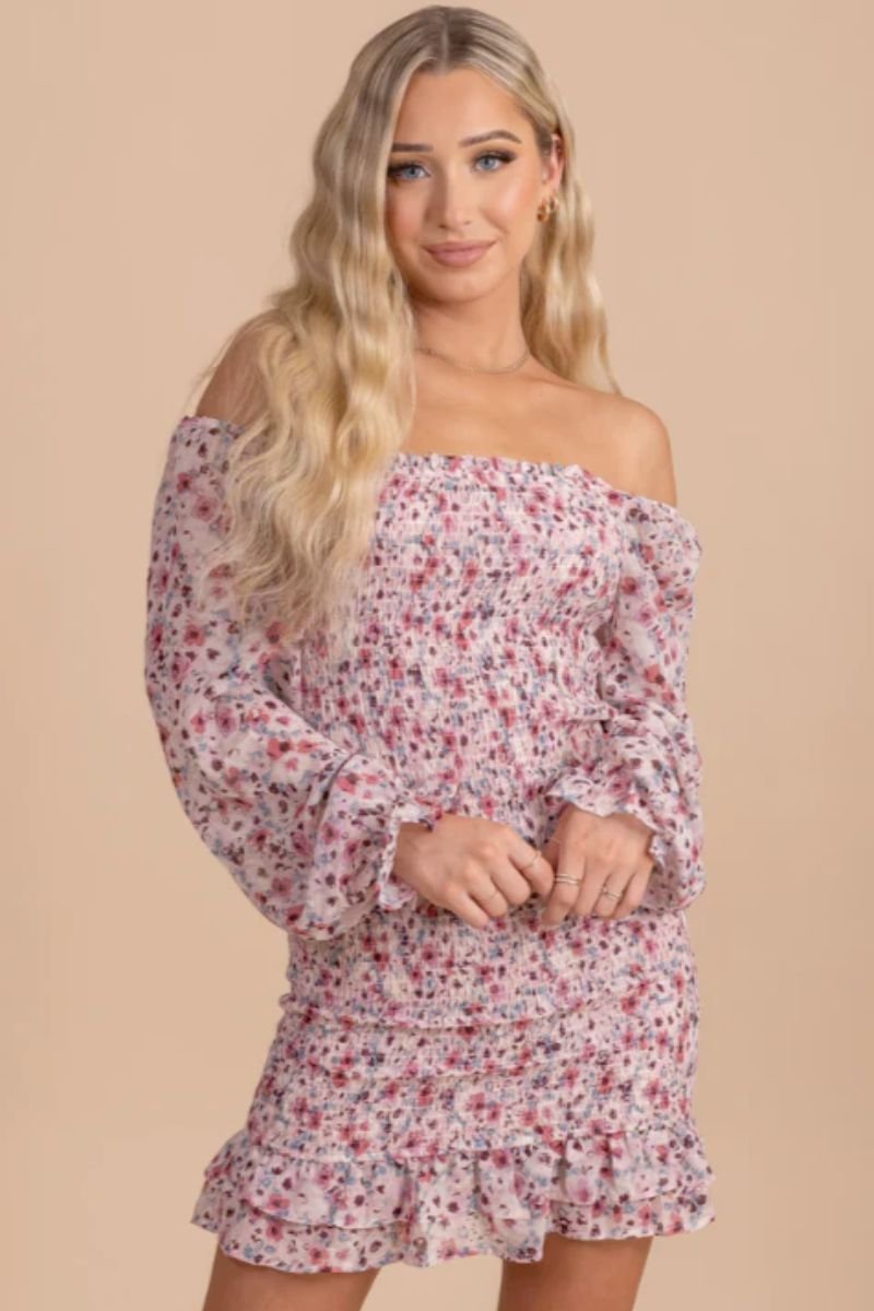 Pace of Nature Smocked Floral Mini Dress. The model is wearing a pink and blue floral form fitting mini dressed with off the shoulder sleeves and a flutter hemline. The model is wearing rings and the sleeves are in a smocked style with a ruffle on the cuff.