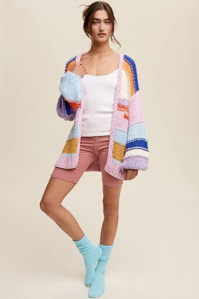 Hand Knit Multi Striped Cardigan. The model is wearing a cardigan with different patches of colors and big sleeves. She is also wearing a white top, pink biker shorts, and blue socks.