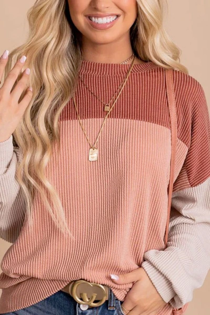 Ribbed color block tunic pullover. The model is wearing a multicolored pink and cream long sleeve top and a belt with gold hardware. She is also wearing gold jewelry, a pink shoulder bag, and blue jeans.