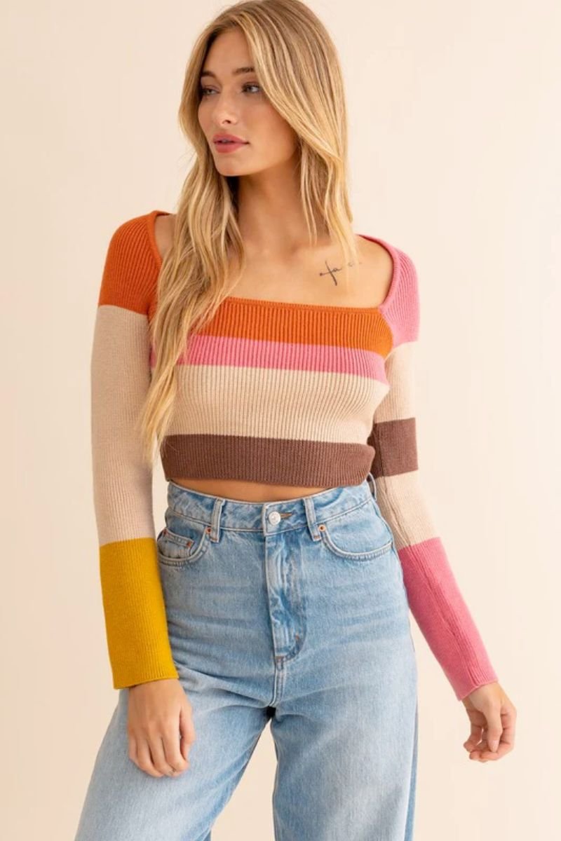 Long Sleeve Color Block Stripe Knit Top. The model is wearing a colorful top with a wide neck line and it is cropped above the waist. She is also wearing blue jeans and has a tattoo near her collar bone.