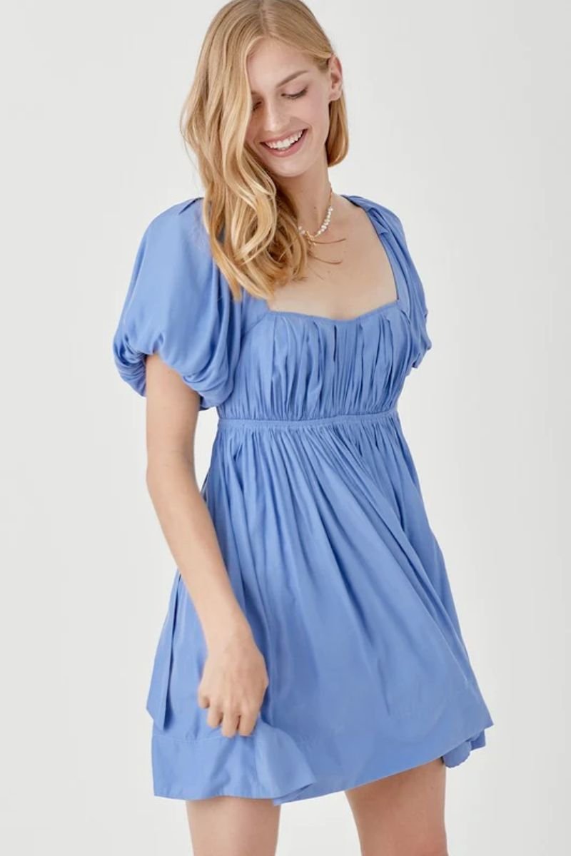 Pleated Front Criss Cross Back Strap Dress. The model is wearing a blue mini dress with a sweetheart neckline and puff sleeves. The model is wearing a necklace with pearls and she had blonde hair.