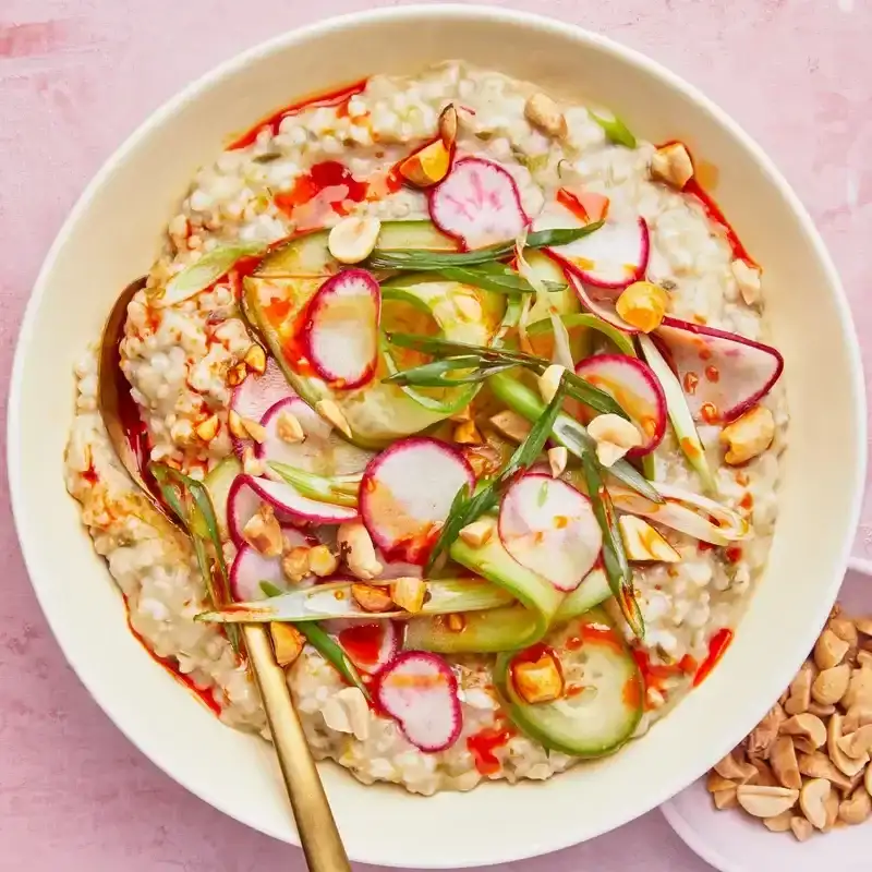 A bowl of savory oatmeal topped with shaved radishes, pickled cucumbers, green onions, and chopped peanuts.