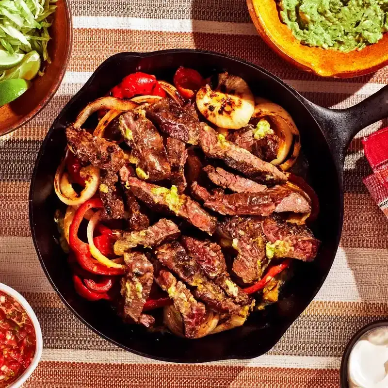Steak fajitas served in a cast-iron skillet next to bowls of salsa, lime wedges, and guacamole.