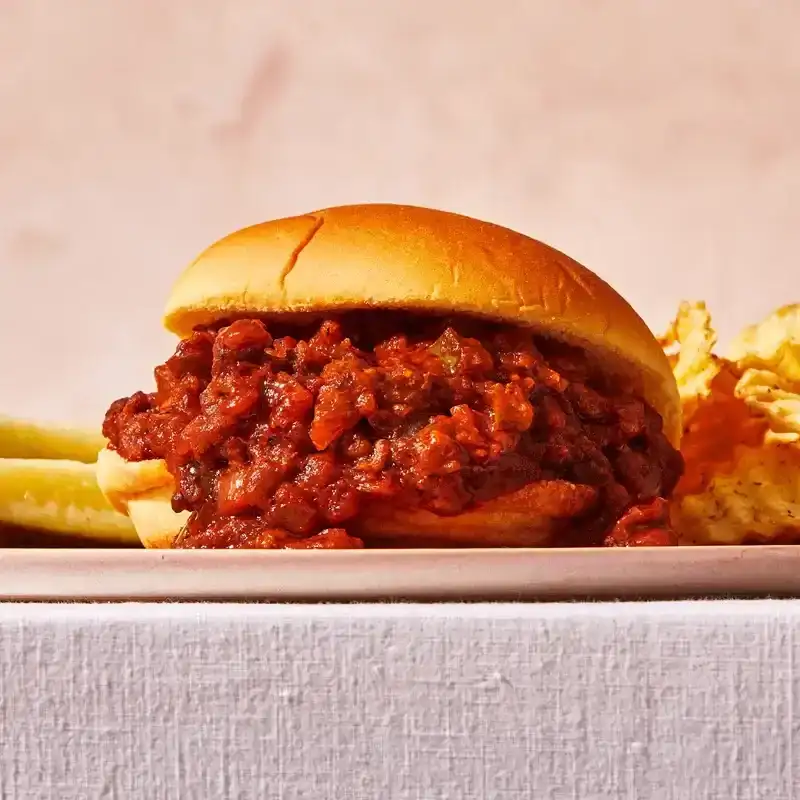 Close-up of a gochujang sloppy joe on a bun, with ridged potato chips and pickles behind it