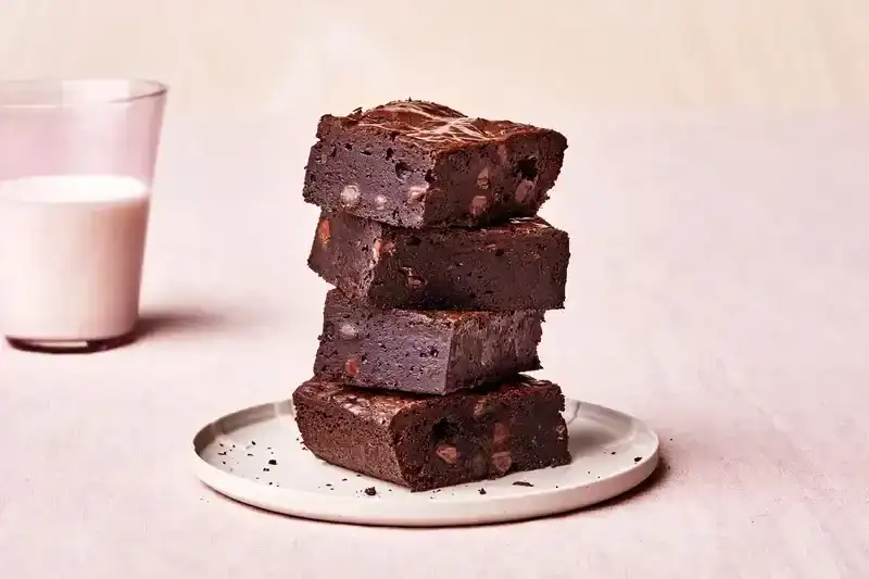 Triple Chocolate Brownies stacked on a white plate with a glass of milk behind it. 