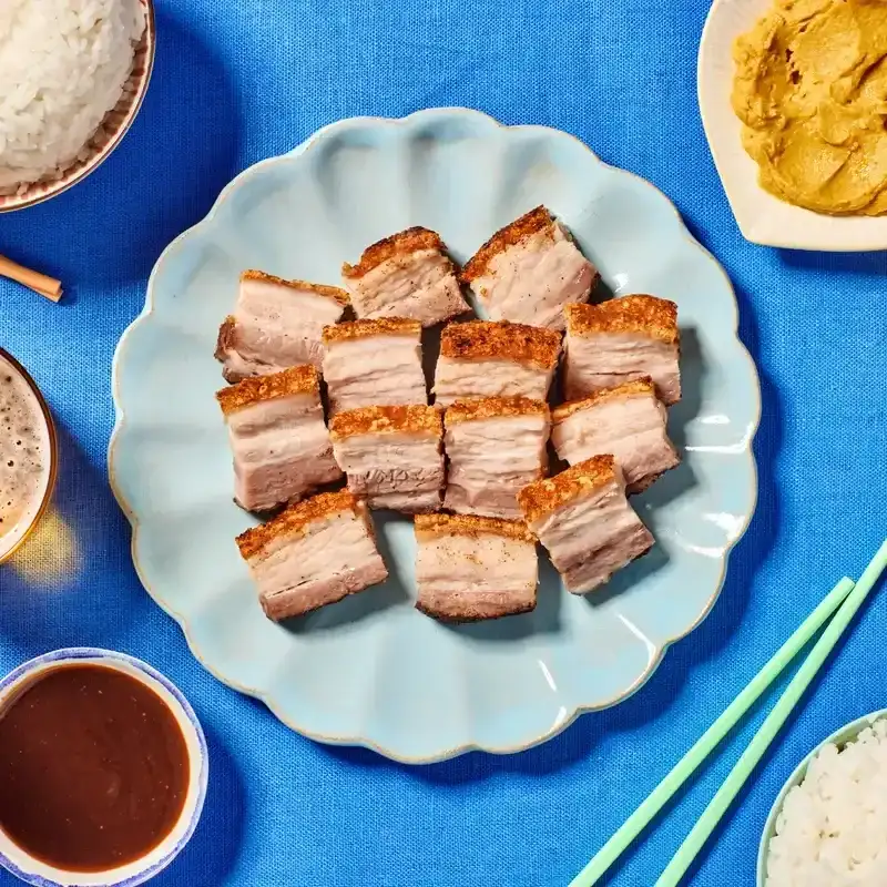 Chopped pieces of cantonese roast pork on a light blue plate next to a variety of toppings on a blue fabric surface.