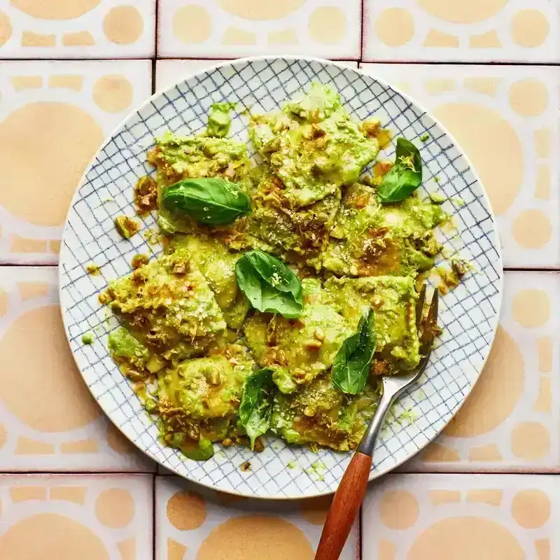 Gridded plate of cheese ravioli covered in green pea sauce, pistachios, and basil, atop pale yellow tiles.