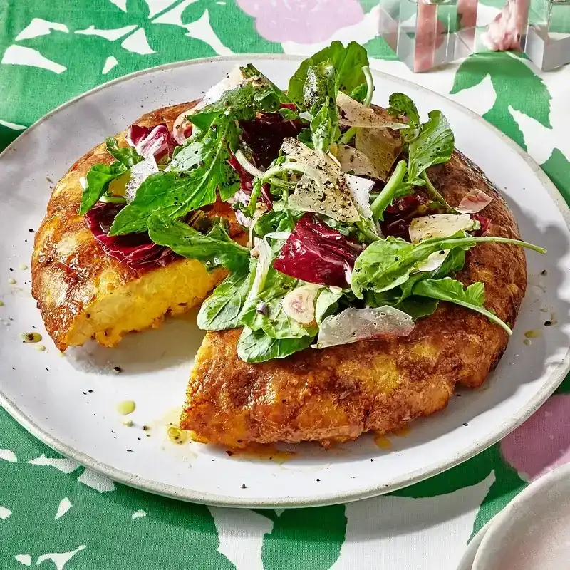 A platter of faux tortilla española, topped with a mixed greens salad and with one wedge cut away.