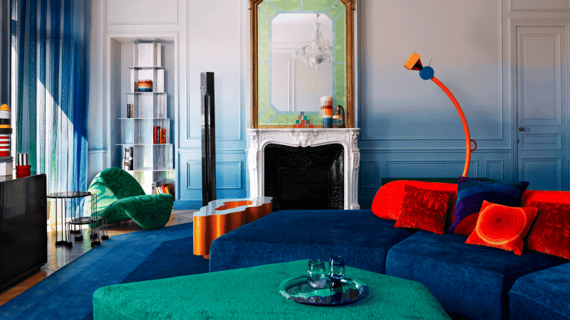 colorful living room with a blue velvet couch, green velvet ottoman, built-in bookshelf, and a antique white fireplace