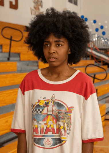 model posing in front of gym bleachers wearing a retro Atari t-shirt and hair in an afro