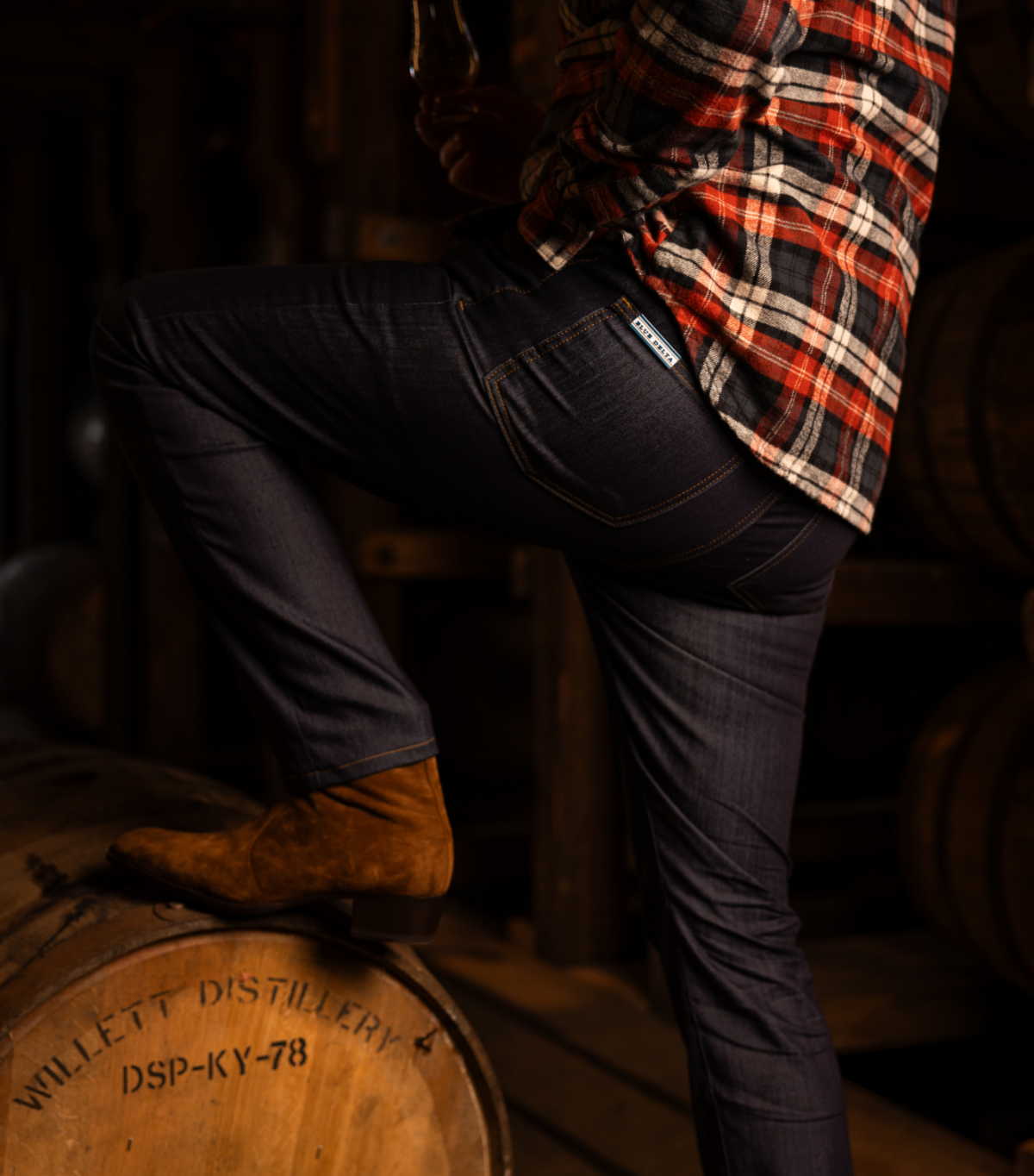 A man is seen from the waist down standing with his left leg up on a cask of whiskey. He wears a red, white, and black flannel shirt and dark jeans from Blue Delta Jeans.