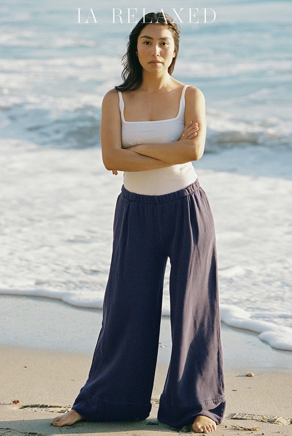 Model stands on beach wearing white camisole and navy fleece wide leg pants.