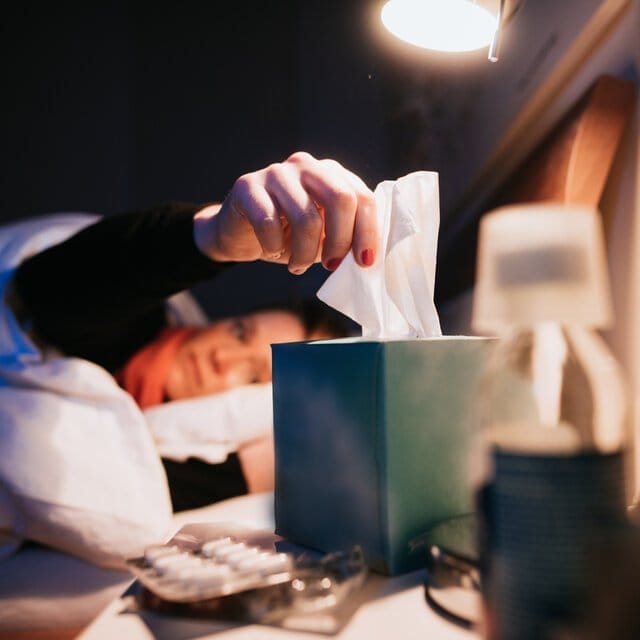 A woman laying in bed grabs a tissue from a box on her bedside table. 