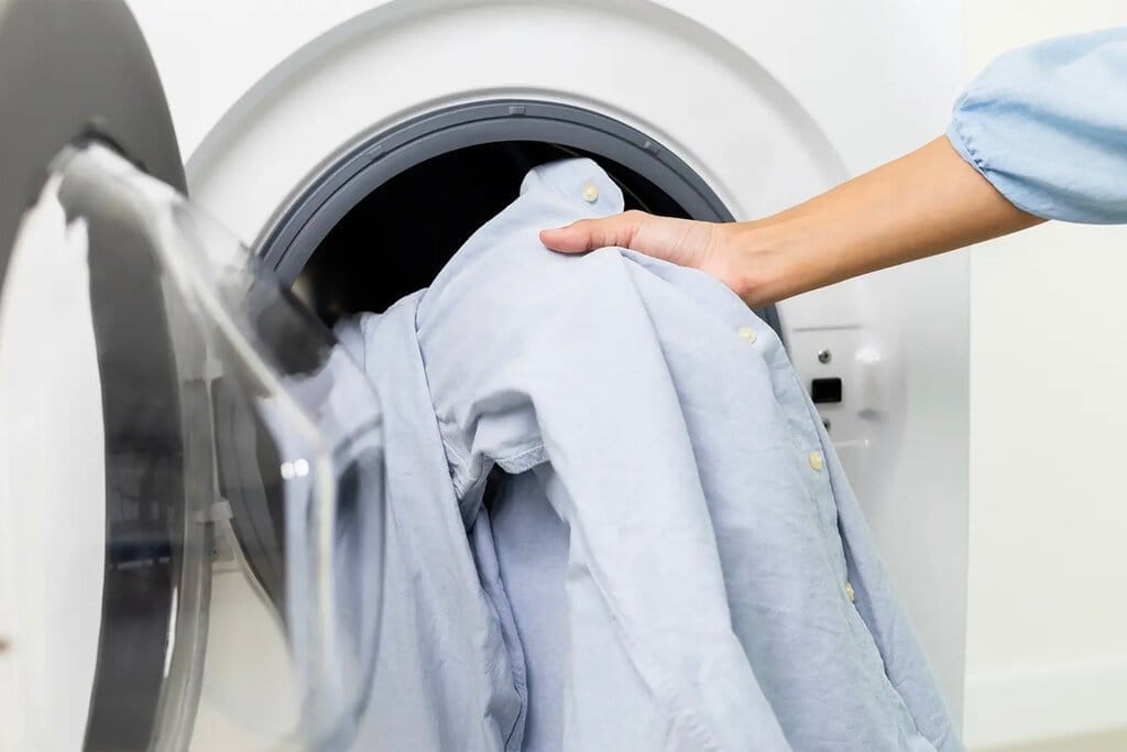 An arm holding a light-blue oxford shirt and putting it in a washing machine.