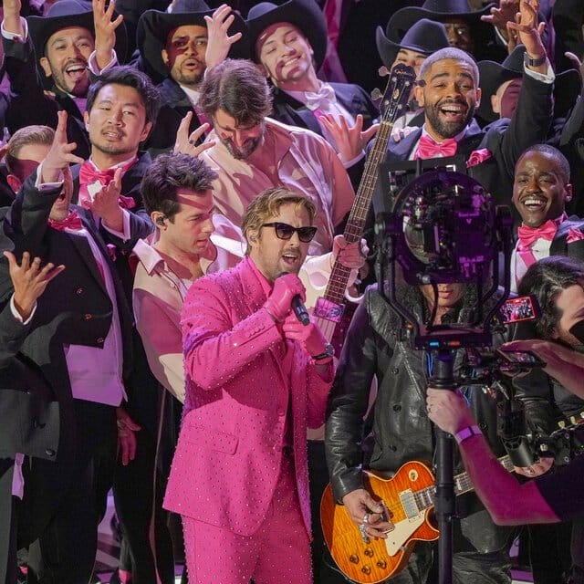 Ryan Gosling, in a pink suit, sings into a microphone. Behind him, a throng of men hold out their arms.