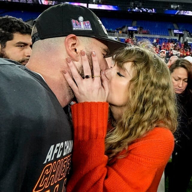 Travis Kelce, left, wearing football pads with an AFC Champion T-shirt and hat that says Super Bowl, kisses Taylor Swift on the field after a game.