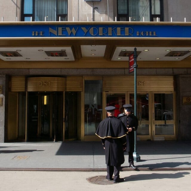 Doormen in uniform stand in the street in front of a marquee reading the New Yorker Hotel.
