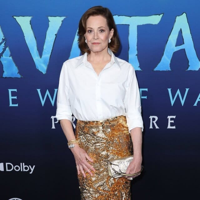 Sigourney Weaver poses in front of a backdrop at an “Avatar” movie event. She wears a button-down white shirt and a long glittery skirt in an abstract bronze and silver pattern. 