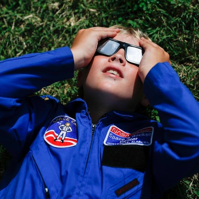 A young boy lying on the grass, with black-framed eclipse glasses on, wears a blue jumpsuit with NASA patches on it. He is pressing the glasses to his face with both hands.
