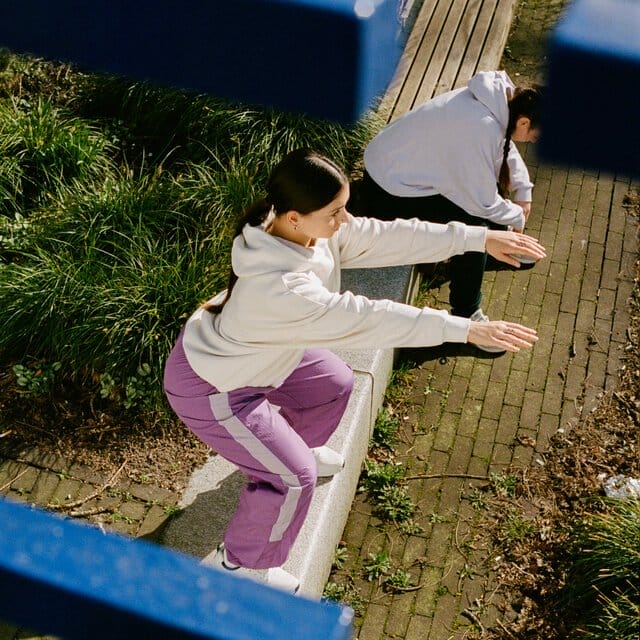 Two women exercise outside on a bench area. One is doing squats while the other leans over on her knees taking a break.