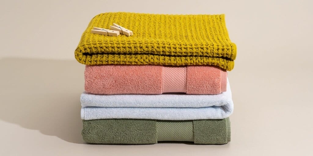 A yellow towel, pink towel, light-blue towel and green towel are folded and stacked on top of one another. Two wooden clothespins are on top of the stack.