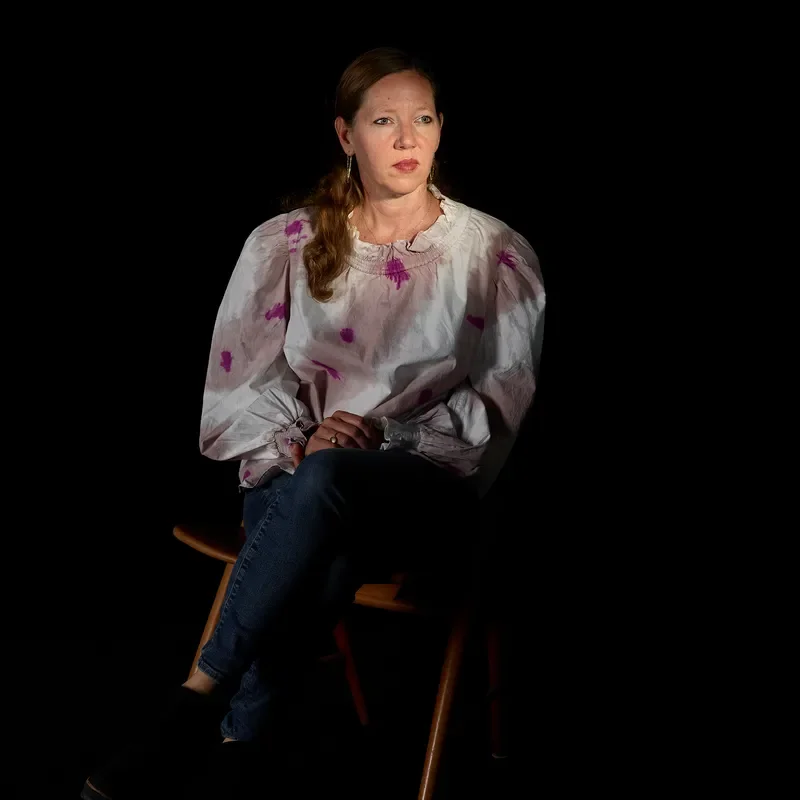 A portrait of Maggie Nelson sitting on a chair in a dark space.