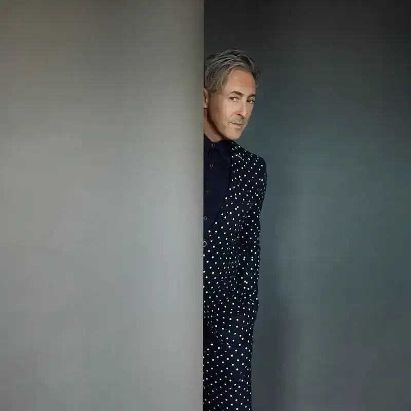Portrait of Alan Cumming peeking around a wall. Cumming is wearing a polka dot suit with shorts and a black button-up shirt. 